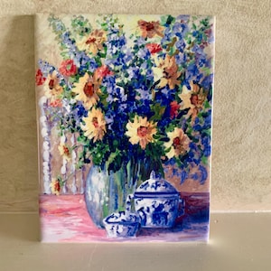 May include: A ceramic tile featuring a floral still life painting. The artwork depicts a vibrant bouquet of sunflowers, blue delphiniums, and red flowers in a blue vase, with blue and white porcelain jars on a pink surface.