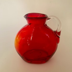 May include: A round, vibrant red glass pitcher featuring a smooth, bulbous body and a clear, curved glass handle attached to the side. The opening at the top is wide with a small spout, and the glass transitions to a subtle orange hue at the base.