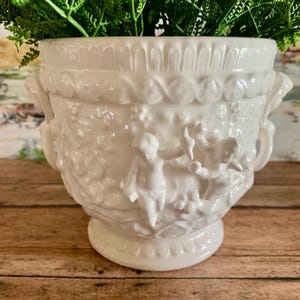 May include: A white ceramic planter with a detailed relief design featuring cherubs and floral elements. The planter has a wide, decorative rim and two side handles. Green foliage fills the top of the planter, creating a contrasting color.