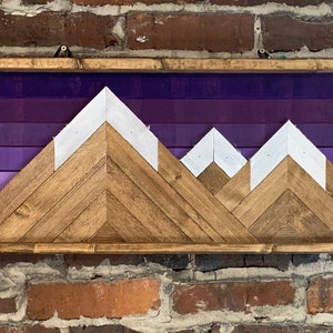 K&ouml;nnte beinhalten: Ein Wandbild aus Holz mit einer Silhouette einer Bergkette vor einem violetten Ombre-Hintergrund. Die Berge bestehen aus Holzbrettern, die in einem Chevron-Muster angeordnet sind, und die Gipfel sind wei&szlig; gestrichen.