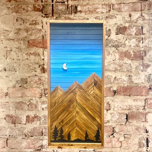 May include: A wooden wall art piece depicting a mountain range with a crescent moon in a blue sky. The mountains are made of brown wood and the sky is painted blue with white stripes. The piece is framed in a dark wood frame.