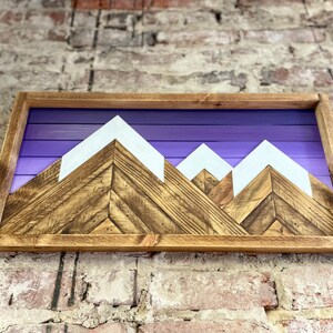 K&ouml;nnte beinhalten: Wandkunst aus Holz mit einer Bergkette vor einem violetten Himmel. Die Berge sind aus Holz mit wei&szlig; bemalten Gipfeln.