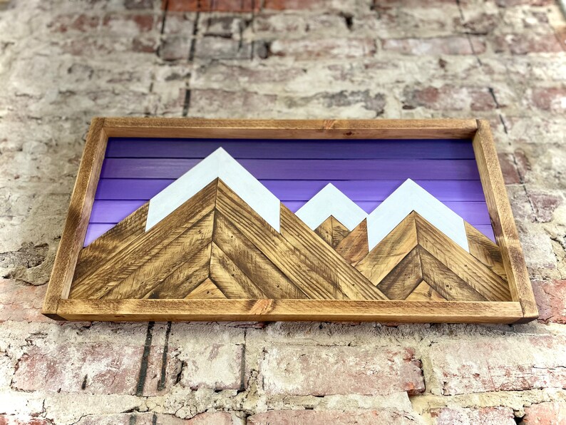 K&ouml;nnte beinhalten: Wandkunst aus Holz mit einer Bergkette vor einem violetten Himmel. Die Berge sind aus Holz mit wei&szlig; bemalten Gipfeln.