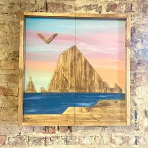 May include: A wooden framed artwork depicting a sunset over a mountain range with a blue ocean in the foreground. The artwork is made of reclaimed wood and painted in shades of pink, orange, blue, and brown.