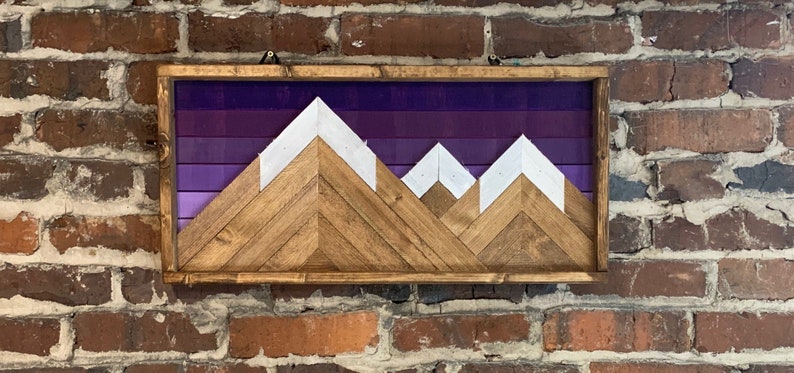 K&ouml;nnte beinhalten: Ein Wandbild aus Holz mit einer Silhouette einer Bergkette vor einem violetten Ombre-Hintergrund. Die Berge bestehen aus Holzbrettern, die in einem Chevron-Muster angeordnet sind, und die Gipfel sind wei&szlig; gestrichen.