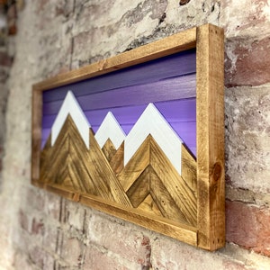 K&ouml;nnte beinhalten: Eine Wandkunst aus Holz mit einer Bergkette und einem violetten Ombre-Hintergrund. Die Berge sind aus Holz mit wei&szlig;en Gipfeln. Die Kunst ist in einem naturbelassenen Holzrahmen gerahmt.