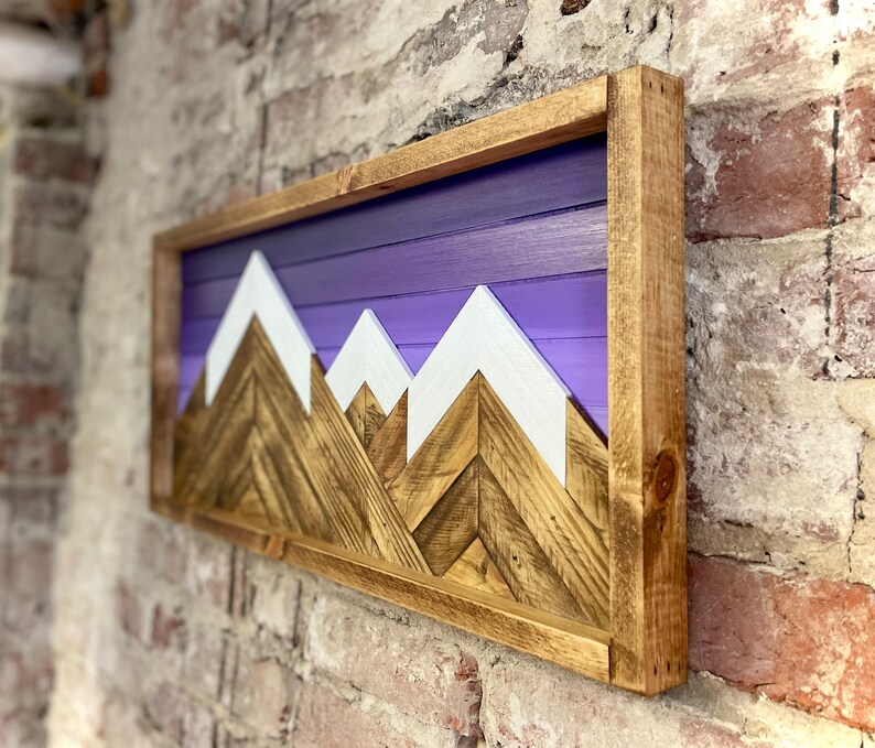 K&ouml;nnte beinhalten: Eine Wandkunst aus Holz mit einer Bergkette und einem violetten Ombre-Hintergrund. Die Berge sind aus Holz mit wei&szlig;en Gipfeln. Die Kunst ist in einem naturbelassenen Holzrahmen gerahmt.