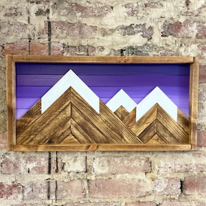 K&ouml;nnte beinhalten: Ein Wandbild aus Holz mit einer Silhouette einer Bergkette mit wei&szlig;en Gipfeln vor einem violetten Verlaufshintergrund. Die Berge sind aus recyceltem Holz gefertigt und in einem dunkelbraunen Holzrahmen gerahmt.