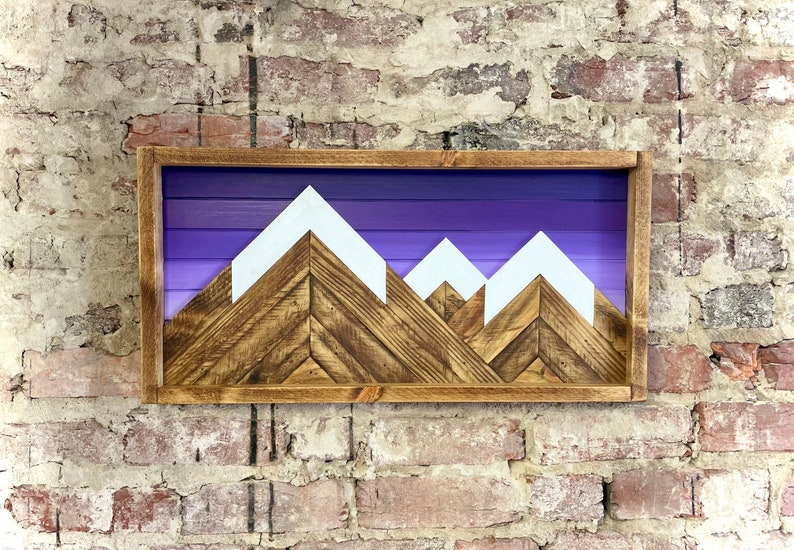 K&ouml;nnte beinhalten: Ein Wandbild aus Holz mit einer Silhouette einer Bergkette mit wei&szlig;en Gipfeln vor einem violetten Verlaufshintergrund. Die Berge sind aus recyceltem Holz gefertigt und in einem dunkelbraunen Holzrahmen gerahmt.