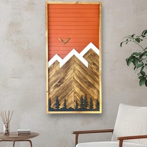 May include: A wooden wall art piece with a mountain scene. The mountains are made of wood and painted white. The background is a red-orange color. The art piece is framed in wood.
