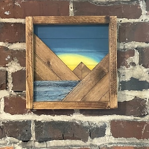 May include: A wooden wall art piece depicting a mountain range with a yellow sun setting behind the peaks. The mountains are made of wood slats painted in shades of blue and brown. The piece is framed in a dark brown wood frame.