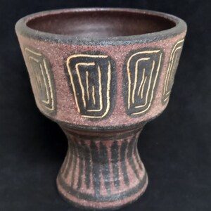 Può includere: Un vaso in ceramica marrone con un design geometrico. Il vaso ha un'ampia apertura e una base stretta. Il design è in nero e oro.
