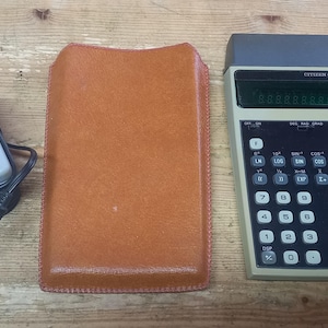 Könnte beinhalten: Ein grau-schwarzer Taschenrechner mit einer braunen Ledertasche und einem grauen Netzteil. Der Rechner hat ein Display, einen Ziffernblock und Funktionstasten. Der Text auf dem Rechner lautet "HITACHI SRI".