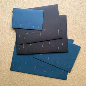 May include: A collection of rectangular envelopes in dark blue and black, each featuring a delicate, hand-drawn pattern of gold and silver stars and celestial sparkles scattered across the lower portion of the front surface of the paper.
