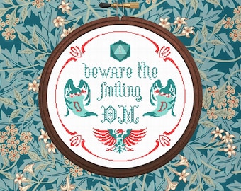 May include: A white embroidery hoop with a red, green, and blue cross-stitch design. The design features a single die with the number one, two winged figures, and the text "Beware the Smiling DM".
