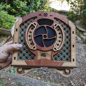 May include: A wooden speaker with a blue and green geometric design. The speaker has a round, black speaker grille with a wooden frame. The speaker has a knob on the top with the symbols + and -.