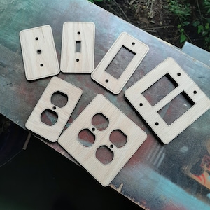 Könnte beinhalten: Sechs hölzerne Wandplattenabdeckungen in verschiedenen Größen und Formen. Die Abdeckungen sind unbehandelt und bereit zum Lackieren oder Beizen. Die Abdeckungen umfassen einen einfachen Lichtschalter, einen doppelten Lichtschalter, eine einzelne Steckdose, eine doppelte Steckdose und zwei einfache Lichtschalter.