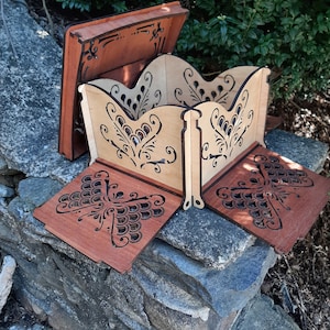 Puede incluir: Una caja de madera con un diseño de mariposa. La caja está hecha de dos tipos diferentes de madera, una clara y otra oscura. La caja está abierta y la tapa está levantada. La caja está sentada sobre una superficie de piedra.