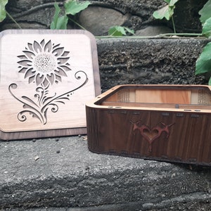 Può includere: Una scatola di legno con un design a girasole sul coperchio e un design a cuore sul fondo. La scatola è aperta e l'interno è visibile.