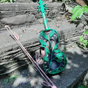 Può includere: Un violino di legno verde con un design floreale e un archetto rosa. Il violino è appoggiato su un gradino di pietra con foglie verdi sullo sfondo.