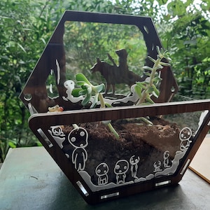 Op de afbeelding: Een zeshoekig terrarium van hout met een transparante glazen voorkant. Het terrarium heeft een silhouet van een persoon die een paard berijdt en verschillende kleine witte figuren. Het terrarium is gevuld met aarde en een kleine vetplant.