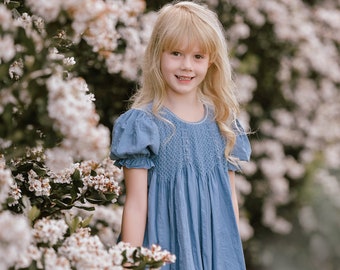 May include: A young girl with blonde hair is wearing a light blue smocked dress and standing in front of a bush of white flowers.