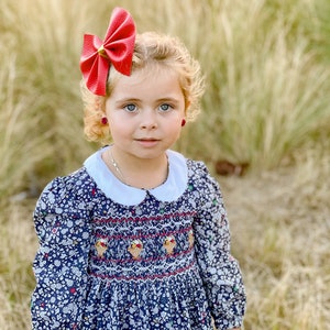May include: A young girl with blonde curly hair wearing a blue dress with a white collar and a red bow in her hair. She is wearing red earrings and has a sweet expression on her face.
