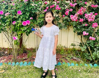 May include: A young girl wearing a white dress with a floral pattern stands in front of a pink rose bush. She is holding a small stuffed koala in her right hand. She is wearing black shoes.
