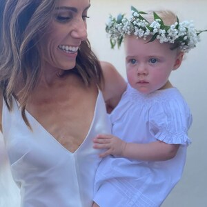 May include: A woman in a white dress smiles while holding a baby girl wearing a white dress and a flower crown.