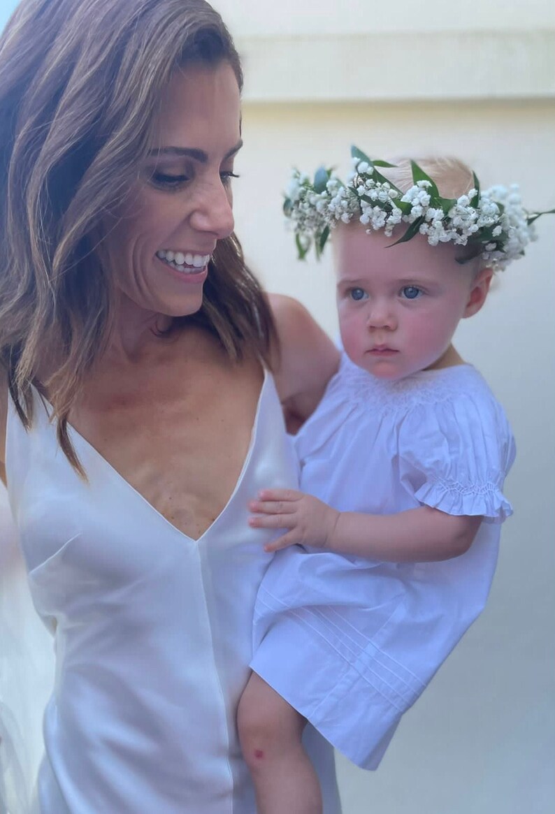 May include: A woman in a white dress smiles while holding a baby girl wearing a white dress and a flower crown.