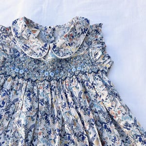 May include: A floral print dress with a Peter Pan collar and smocked bodice. The dress features a white collar trim and small blue floral accents. The fabric has a pattern of blue, yellow, and green leaves and flowers.