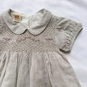 May include: A light beige baby dress with a Peter Pan collar and short, puffed sleeves. The bodice features intricate smocking and delicate pink rose embroidery. The dress has a soft, textured appearance and a tag that reads "Mini Sonata handmade with love."