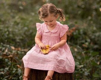 May include: A young girl wearing a pink dress sits on a tree stump in a woodland setting. She is holding a small yellow chick in her hands.