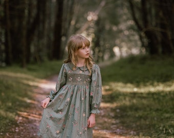 May include: A young girl wearing a green floral dress with puffed sleeves and a white collar stands in a forest setting. The dress has a smocked bodice and a gathered skirt.