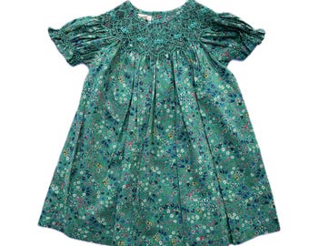 May include: A green floral print dress with short sleeves. The dress has a smocked bodice and a flared skirt. The fabric is a light green with a pattern of small flowers in shades of blue, pink, and white. The dress is designed for children.
