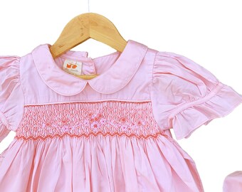 May include: A pink smocked dress with a white collar and short puffed sleeves. The dress features a floral embroidery design and a label that reads "Miss Lovely" on the front.