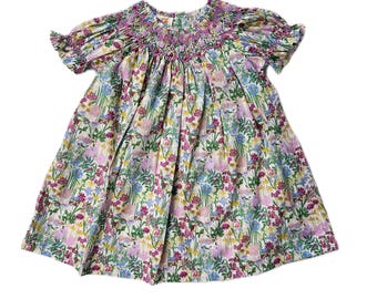 May include: A white smocked dress with a floral print featuring pink, blue, yellow, and green flowers. The dress has short puffed sleeves and a round neckline.