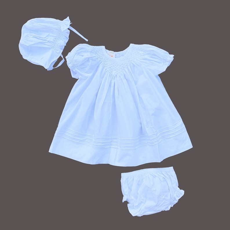 May include: A white baby dress with puffed sleeves, a smocked bodice, and a matching bonnet. The dress has a ruffled hem and is made of a soft, lightweight fabric.