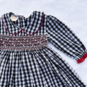 May include: A black and white gingham dress with long sleeves. The dress has a red-trimmed Peter Pan collar and cuffs. The bodice features a smocked design with red and white detailing. The dress is made of a lightweight fabric.
