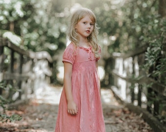 May include: A young girl with long blonde hair wearing a pink dress stands on a wooden bridge. The dress features short sleeves, decorative buttons, and a detailed bodice. The background is blurred with greenery and sunlight.