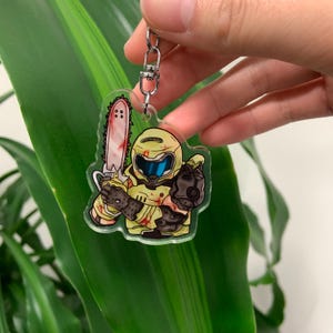 May include: A clear acrylic keyring featuring a cartoon character in a yellow suit and helmet, holding a bloody chainsaw. The character is pointing with a gloved hand. The keyring has a silver metal chain.