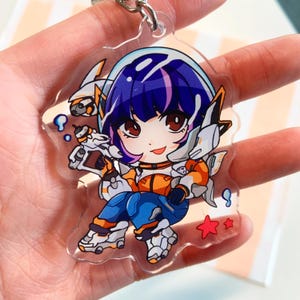 May include: A clear acrylic keychain featuring a chibi-style character with dark purple hair, wearing an orange and blue futuristic suit. The character holds a white and orange blaster, surrounded by small blue bubbles and two red star icons.
