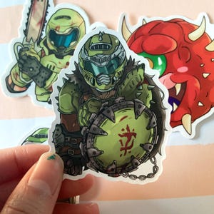 May include: A collection of colorful stickers featuring characters from the video game Doom. The stickers include a green armored character with a shield, a character with a chainsaw, and a red monster with horns.