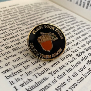 May include: A gold and black enamel pin with the text "Plant Your Trees, Watch Them Grow". The pin features a cartoon acorn with a brown cap and a gold nut.