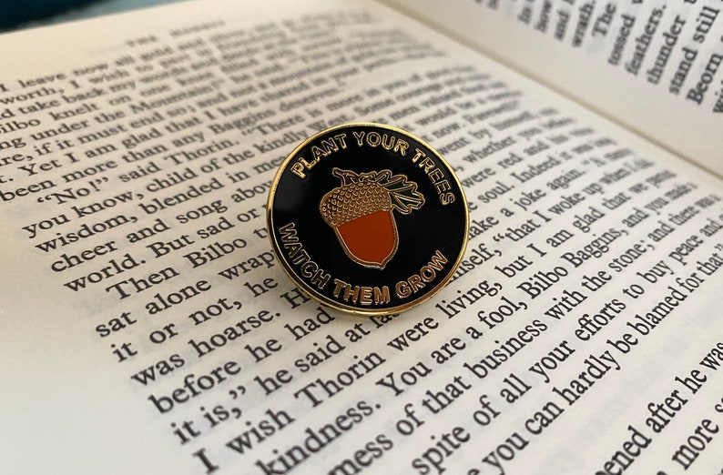 May include: A gold and black enamel pin with the text "Plant Your Trees, Watch Them Grow". The pin features a cartoon acorn with a brown cap and a gold nut.
