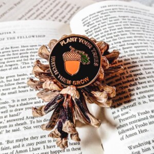 May include: A black enamel pin with a gold acorn and leaves. The pin reads "Plant Your Trees Watch Them Grow".
