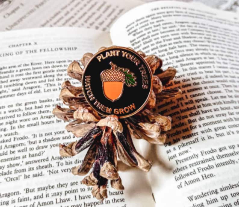 May include: A black enamel pin with a gold acorn and leaves. The pin reads "Plant Your Trees Watch Them Grow".