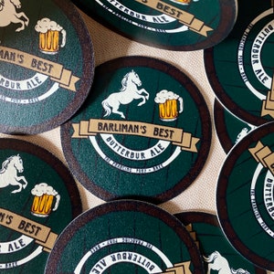 May include: A set of six round green coasters with a brown wooden barrel design. Each coaster features a white horse, a glass of beer, and the text "Barliman's Best Butterbur Ale The Prancing Pony - Bree".