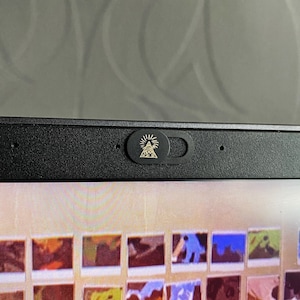 以下が含まれることがあります： 黒いウェブカメラカバー。金色のデザインで、目と太陽光線のある三角形が特徴です。カバーは黒いノートパソコンの画面上部に取り付けられています。