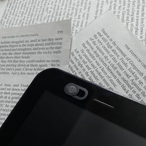 May include: A black e-reader with a black and silver webcam cover featuring a stylized design of a diamond with a crown on top. The e-reader is lying on top of a page of a book.
